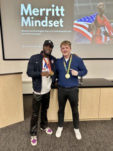 Two men wearing Olympic gold medals standing next to each other in front of a large screen with a presentation displayed on it.