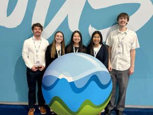 Five college students standing together, smiling, in front of a light blue wall with white script and behind a ball with colorful blue and green waves on it.