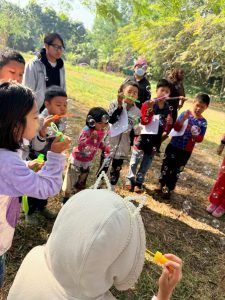 Several children blowing bubbles in a park-like setting with grass and trees