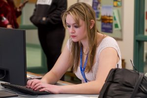 Woman with long blonde hair sitting at a table looking at a computer monitor typing on a keyboard.
