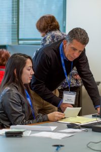 Man wearing a dark sweater and a lanyard around his neck looking at a form with a woman who is seated at a table in front of a computer