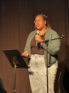 African American woman standing behind a music stand holding a microphone speaking to an audience