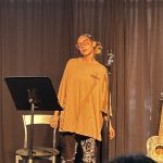 Female singer wearing glasses with her hair pulled back performing on a stage in front of an audience
