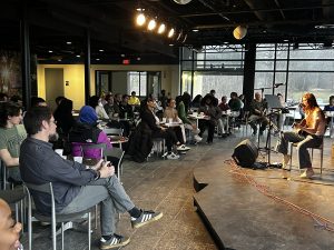 A musician seated on a stage in front of a room full of people seated at tables listening to her performance. 