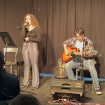 Female singer with long curly hair standing, singing with a male guitar player seated beside her playing the guitar.