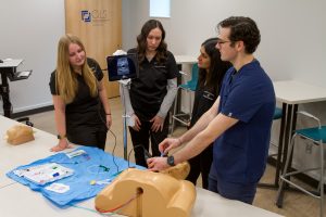 Group of PA students gathered around a manikin while an instructor demonstrates a procedure
