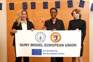 Three women standing together holding a white banner with black text and round logos on it