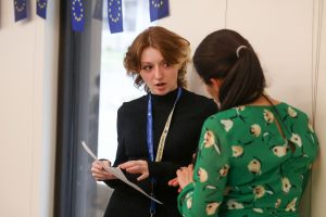 Two women standing, facing each other and talking, one is wearing a green dress and one is wearing a black dress.