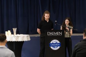 A female college student speaks into a microphone talking to an audience while a male college student stands next to her behind a podium.