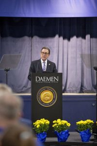 Man wearing glasses, a dark gray suit, white shirt and a purple paisley tie standing behind a podium speaking to an audience