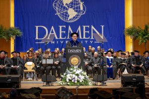 University President wearing cap and gown, standing behind a podium speaking to a crowd with other men and women seated behind him wearing caps and gowns