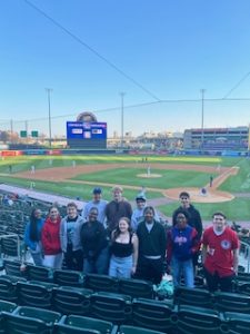 Group of college students standing together in the stands of a minor league baseball stadium with the field showing behind them.