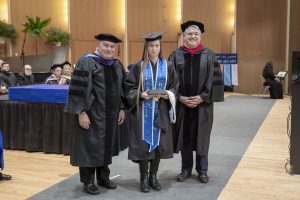 Female college student wearing a cap and gown, standing between two men also wearing caps and gowns