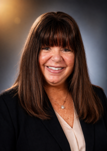 Woman with long brown hair and bangs, wearing a light blouse and a dark blazer smiling with a blueish-brown backdrop behind her