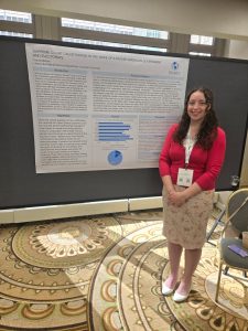 Female college student dressed in a khaki skirt and red sweater standing next to a large poster with data on it on an easel