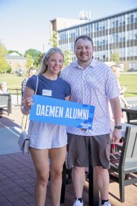 A man and woman standing outside on a college campus holding a Daemen Alumni sign