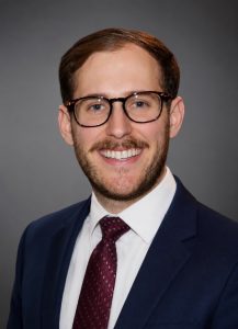 Headshot of a man with a mustache wearing glasses and a suit and tie smiling in front of a gray backdrop