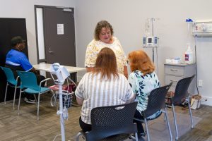 Simulated hospital waiting room, with a man seated at a table and a women interacting with two other women who are seated