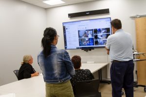 A meeting room with a large monitor on the wall displaying various screens while four people gathered around a long table observe the videos