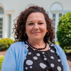 Woman with brown curly hair wearing a black polka dot shirt and light blue cardigan sweater standing in front of a white building