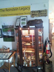 Old windowed cabinet with artifacts on the shelves and photos and signs hanging on the wall