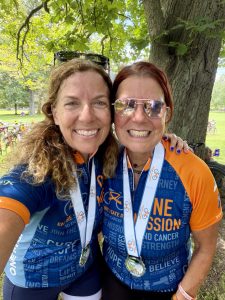 Two smiling women standing arm in arm wearing matching cycling jerseys and medals around their necks