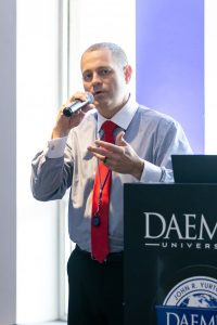 Man wearing a dress shirt and w red tie holding a microphone speaking to an audience from a podium with the Daemen University logo on it