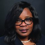 Headshot of an African American woman wearing glasses and a black blouse in front of a dark gray background