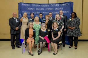 Group of people, some standing, some sitting, in front of a blue step and repeat backdrop with white Daemen letters