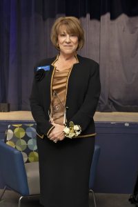 Woman wearing a black suit and shimmery top standing in front of a stage