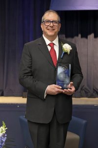 Man wearing a dark suit and red tie holding a trophy standing in front of a stage