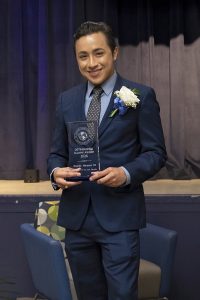 Man wearing a navy suit, blue shirt and dark tie holding a trophy standing in front of a stage
