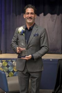 Man wearing a gray suit and dark shirt holding a trophy standing in front of a stage
