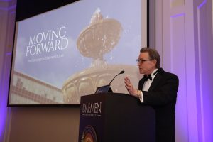 Man wearing a tux standing behind a podium next to a large screen with a photo and text overlay displayed on it