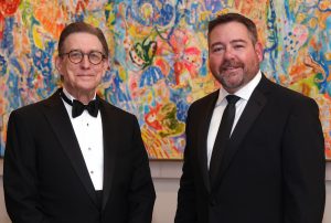 Two men dressed in black tie attire standing in front of a colorful painting