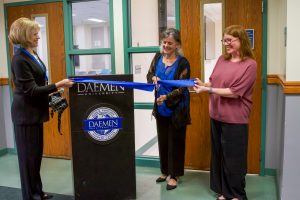 Two women holding a blue ribbon and another woman cutting the ribbon with an oversized pair of scissors