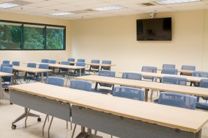 Large classroom with tables, chairs, windows and a wall-mounted TV