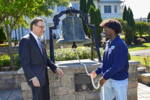 President Olson standing with a male college student in front of Founders Bell on Daemen's campus.