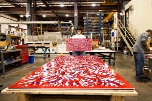 Large metal red, white and blue artwork on a table with a woman standing at the end of the table holding a smaller replica