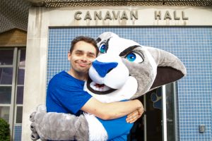 A man hugging the Daemen Mascot, Willie, in front of Canavan Hall