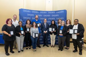 A group of people standing with President Gary A. Olson in front of a blue backdrop with white Daemen University text repeated on it