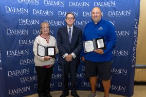 Two people standing with President Gary A. Olson in front of a blue backdrop with white Daemen University text repeated on it