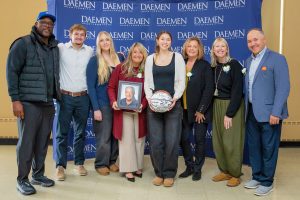 Men and women standing together in front of a blue Daemen step-and-repeat backdrop holding a framed picture of former basketball coach and holding a basketball