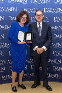 A woman standing with President Gary A. Olson in front of a blue backdrop with white Daemen University text repeated on it