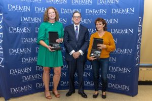 Two women standing with President Gary A. Olson in front of a blue backdrop with white Daemen University text repeated on it