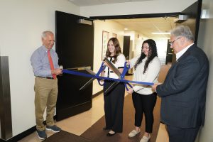 Two men holding oversized ribbon while two female college students cut the ribbon with oversized scissors