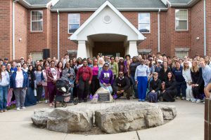 Large group of people standing together around an engraved rock in front of a brick building