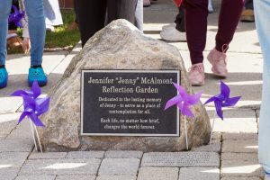 Large stone with an engraved metal plaque attached with purple pinwheels in the ground around it